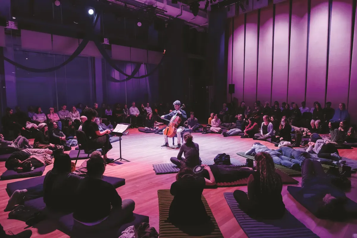 Intimate concert performance with a cellist under violet stage lighting, audience seated on floor cushions