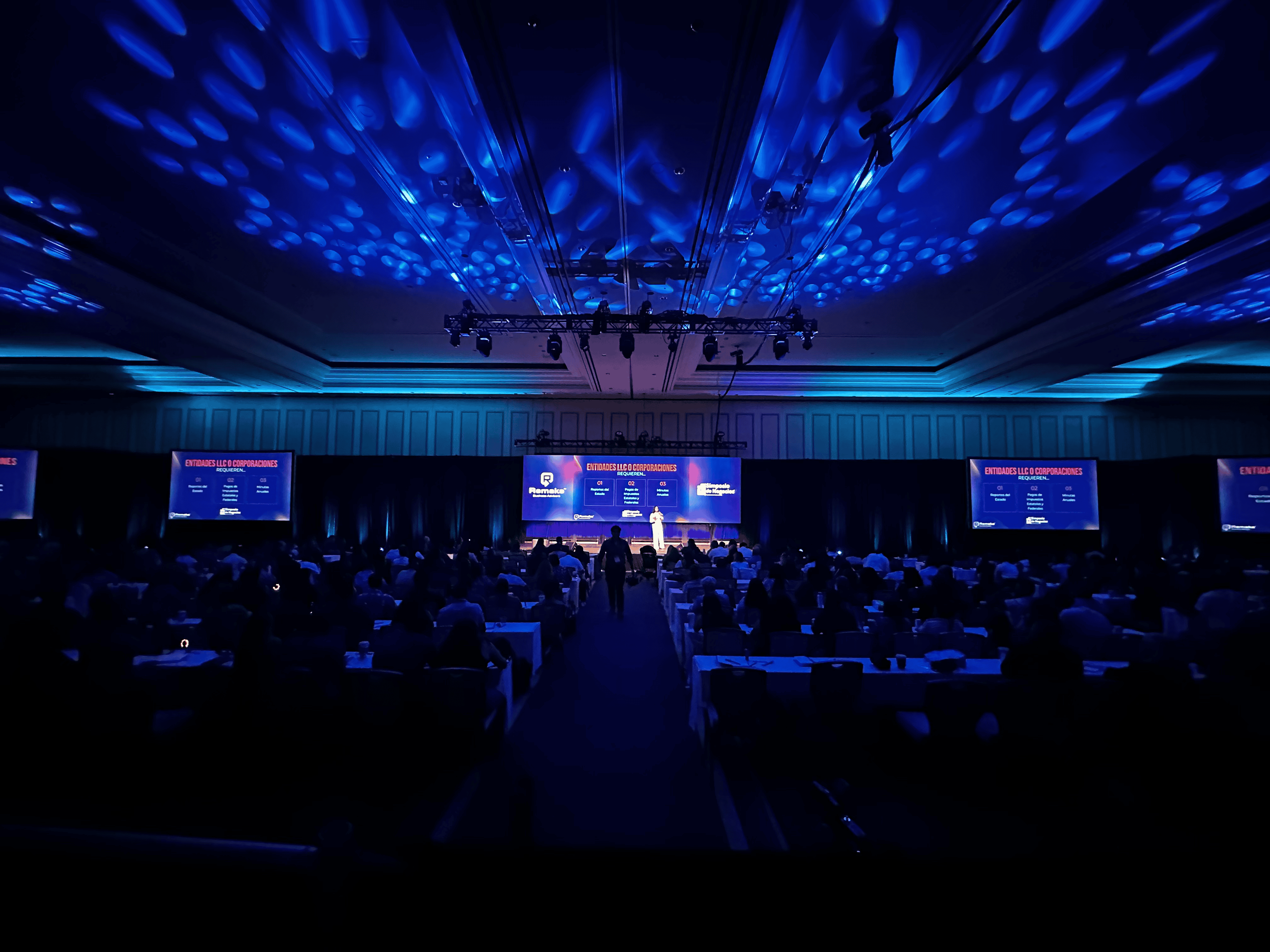 Large corporate conference ballroom with a truss-rigged stage, presentation screens, and blue ambient lighting