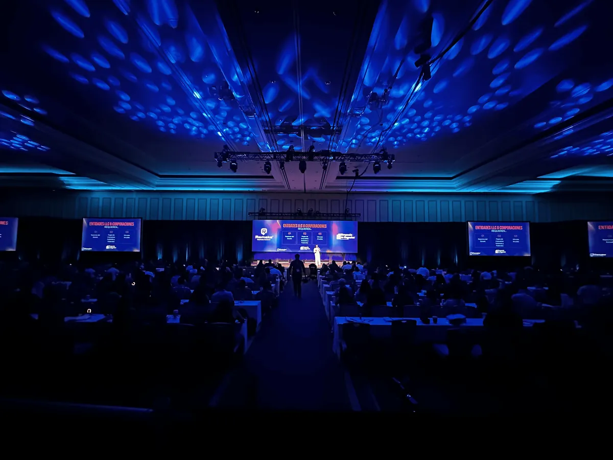 Large corporate conference ballroom with a truss-rigged stage, presentation screens, and blue ambient lighting