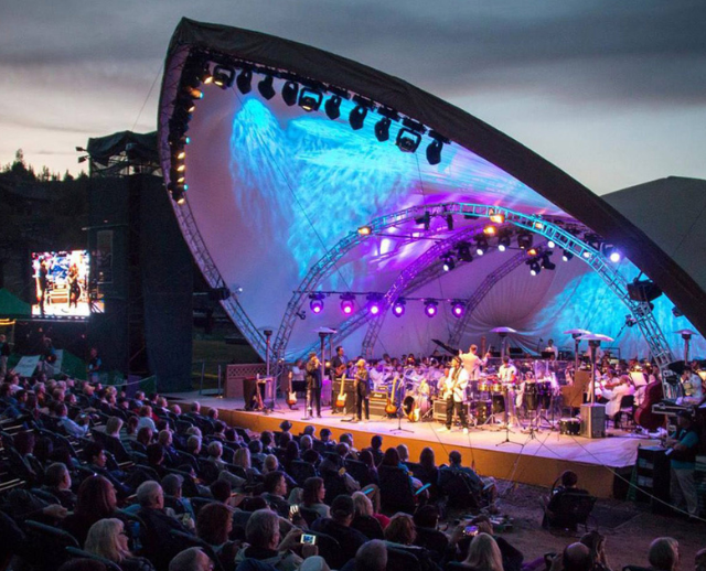 Large outdoor amphitheater concert at dusk with a truss-rigged stage canopy lit in blue and violet