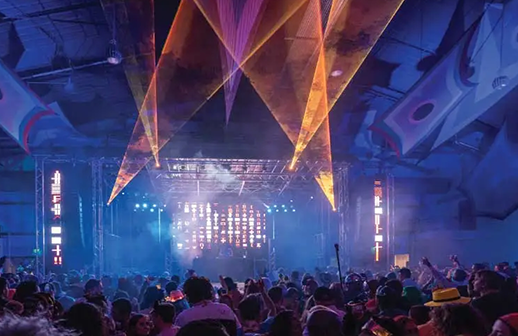 Warehouse rave with truss-rigged stage lighting and amber and violet laser beams over the crowd